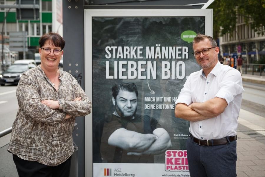 Auf zahlreichen City-Light-Säulen wirbt die ASZ Heidelberg für einen bewussteren Umgang mit dem Bioabfall. Sylvia Hafner, Leiterin der ASZ Heidelberg, und Umweltbürgermeister Raoul Schmidt-Lamontain präsentieren exemplarisch bei einem Fototermin an der Haltestelle Seegarten eines der verschiedenen Motive der Informationskampagne. (Foto: Stadt HEedelberg) Sylvia Hafner und Umweltbürgermeister Raoul Schmidt-Lamontain stehen vor der City-Light-Säule mit dem Aufdruck: "Starke Männer Lieben Bio", begleitet wird der Aufdruck von dem Bild eines Arme verschränkenden, stark aussehenden Mannes.