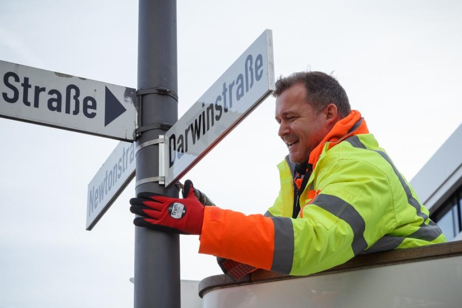 Von der Schlosserei bis an den Schildermast: Bürgermeister Raoul Schmidt-Lamontain begleitete beim Termin die Mitarbeitenden der ASZ Heidelberg bei der Fertigung und Montage von Straßenschildern. In rund drei Metern Höhe wurden die Schilder schließlich an mehreren Stellen in der Bahnstadt montiert. (Foto: Stadt Heidelberg) Zu sehen ist ein Mann in gelber und orangener Arbeitskleidung, der gerade ein Straßenschild anbringt.